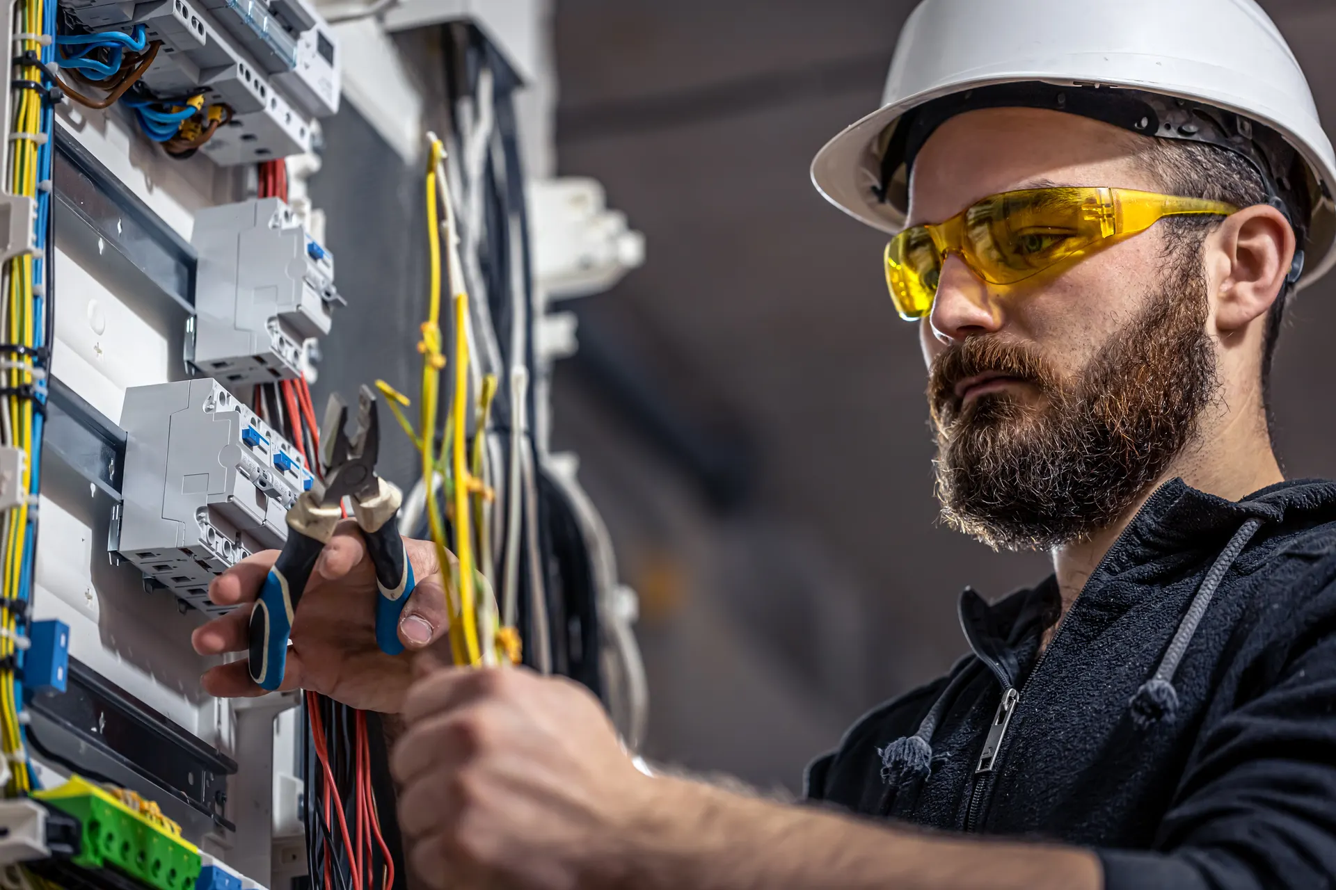 Eletricista com capacete e óculos de proteção trabalhando em quadro de distribuição elétrica